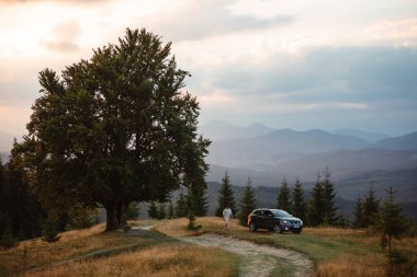 A young man near car. The car is parked near a big old beech tree in the Carpathians