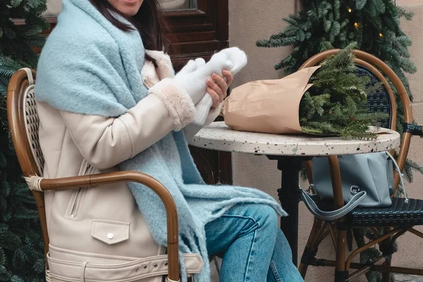 beautiful fashionable woman sitting on the chair at outdoors cafe with christmas tree bouquet blue hat white coat