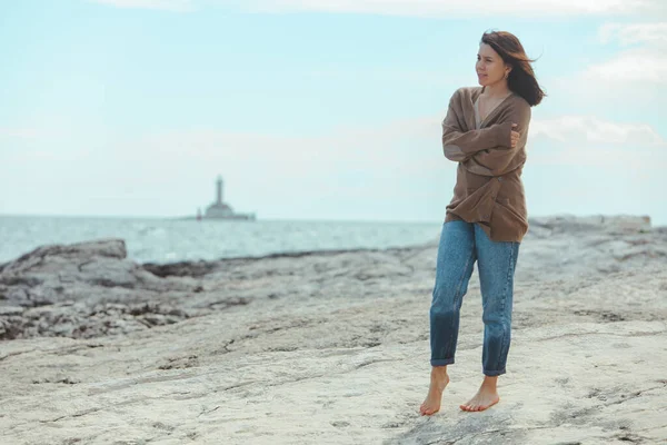 woman walking by rocky sea beach in wet jeans lighthouse on background. windy weather. summer vacation. carefree concept