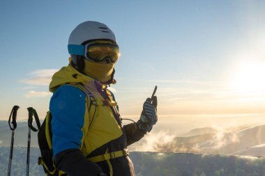 Kadın kayakçı telefonda dağların tepesinde gün batımının fotoğrafını çekiyor.