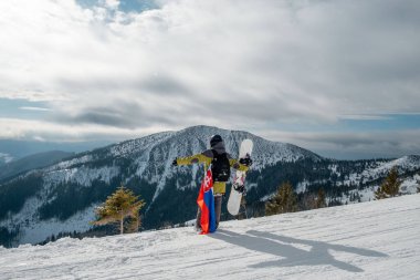 Slovakya bayrağı taşıyan snowboardcu kayak merkezinin yamacında arka planda güzel dağlar var.