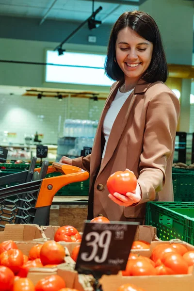 一个千禧年女人用推车在超市里买食物的画像 一位女顾客选择西红柿 从商店的货架上取货 — 图库照片