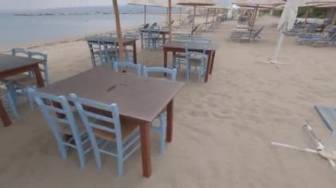 Bulutlu bir akşamda boş bir restoran ve plaj barı hipervane oldu. POV hızı, Yunanistan 'da kumlu bir sahilde, mavi sandalyeli ve deniz kenarında güneşlenen ahşap masalarda yürüyor..