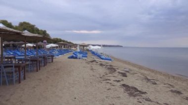 Bulutlu bir akşamda boş bir restoran ve sahil barı. Taverna ahşap masalar mavi sandalyeler ve deniz kenarında güneşlenme yatakları Yunanistan 'da kumlu bir sahil kenarında.
