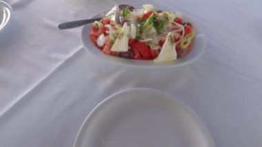 Yunan salatasını peynirli, soğanlı, domatesli, maydanozlu ve salatalıklı servis et. El, beyaz bir tabak taverna masasında, ekmek dilimlerinin yanında bir kaşık ve çatal xoriatiki ile servis ediyor..