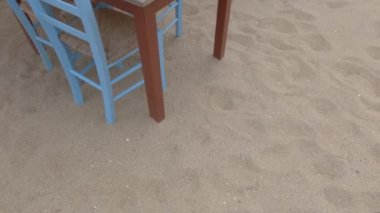 Bulutlu bir akşamda deniz kenarında boş bir restoran. Yunanistan 'da kumsalda mavi sandalyeli taverna ahşap masaları.