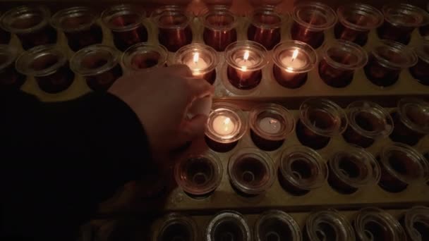 カトリック教会のスタンドでお祈りのワックスキャンドルを点灯手 暗い背景を背景に穏やかな炎を持つ金属製の容器にキリスト教の祈りのために使用される光と光のない白い視覚的なキャンドル — ストック動画