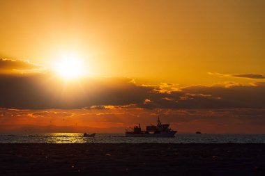 Turuncu gün batımından önce balıkçı teknelerinin akşam manzarası. Akdeniz kıyılarına yakın seyreden gemiler alçak bulutlu parlak gökyüzüne karşı.