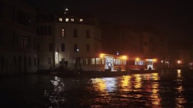 Venedik İtalya ACTV Vaporetto kamu su otobüsünden gece uzaklaşırken, küçük ahşap tekne arka planda aydınlık geleneksel binalarla Büyük Kanal 'ın sakin sularını geçiyor..