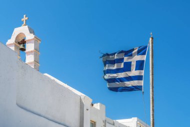 Beyaz boyalı bir Ortodoks kilisesinin önünde Mykonos Adası 'nda çan kulesi ve Hristiyan haçı ile güçlü bir rüzgârın esmesi gereken bir Yunan bayrağı dalgalanmaktadır..