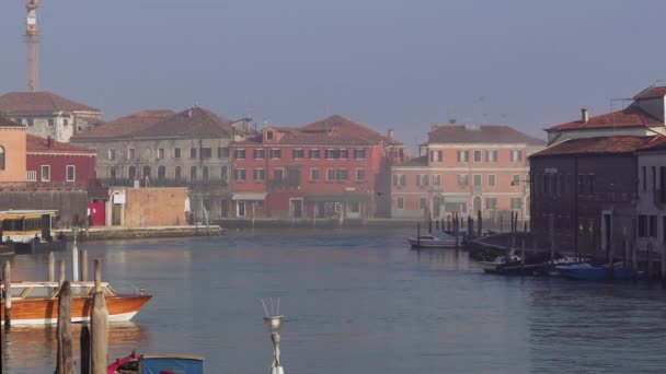 ヴェネツィアイタリアムラーノ島の日の景色いくつかの霧の下で伝統的な低上昇の建物と運河の穏やかな海に沿って木製の棒に係留ボート — ストック動画