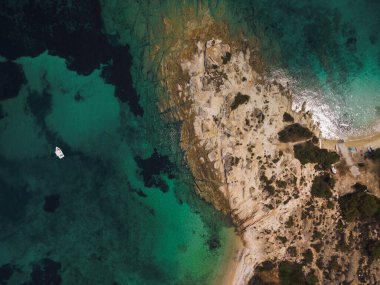 Yunanistan 'ın Chalkidiki yarımadasındaki Akdeniz' de yeşil plantasyonu olan kayalık sahil etrafındaki sakin sulardaki teknenin insansız hava aracı görüntüsü