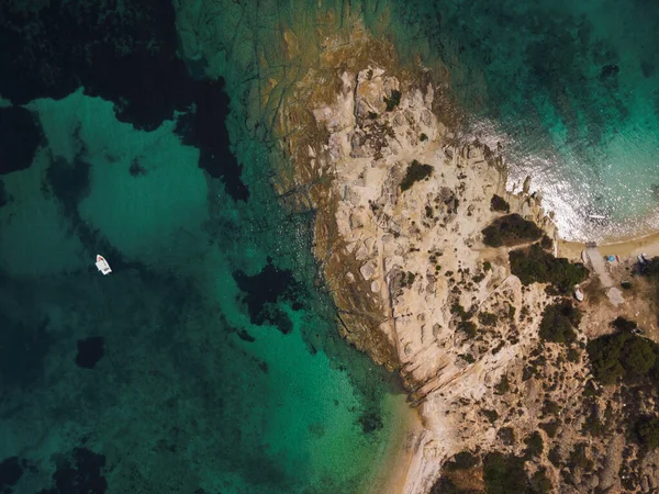 Yunanistan 'ın Chalkidiki yarımadasındaki Akdeniz' de yeşil plantasyonu olan kayalık sahil etrafındaki sakin sulardaki teknenin insansız hava aracı görüntüsü