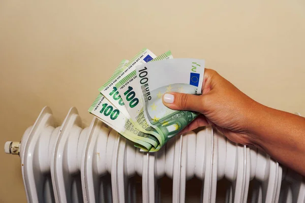 Home winter heating prices going up concept. Female trying to get warm against a white radiator, holding euro paper money.