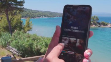 Euboea, Yunanistan - 3 Temmuz 2022: Disney + Service VoD içeriğiyle kaydırma ekranı. Elinde cep telefonu olan bir kadın, plaj arka planında görüntüler yayınlayan sağlayıcı sayfası.