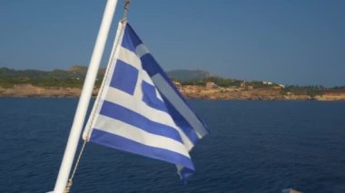 Day sea view of real Greek flag with cross, blue and white stripes waiving on the must of a sailing ship close to the coast in Greece, against blue sky.