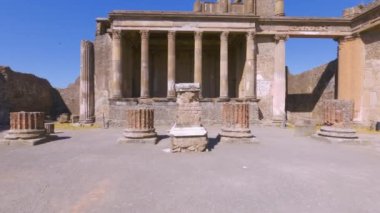 Archaeological area of Pompeii, Italy POV walking on Roman architecture empty basilica monument area with Corinthian colonnades and Ionic capitals against blue sky.
