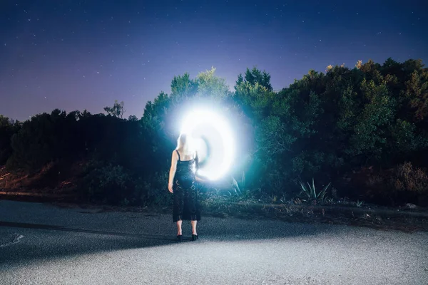 Long exposure of female drawing a portal shaped spiral with a bright light source before a starry night at a deserted rural scenery.