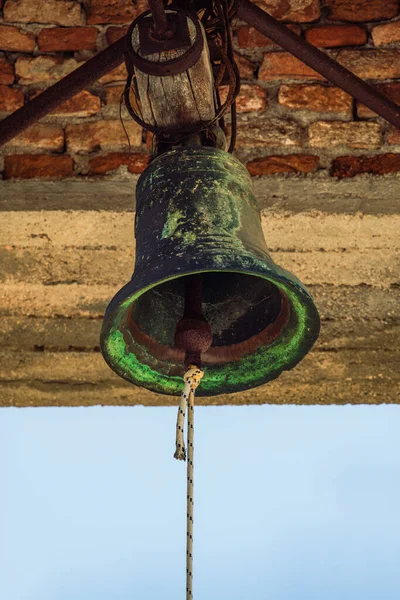 Rusty church old bronze bell hanging from a wooden poll against a brick wall bell tower.