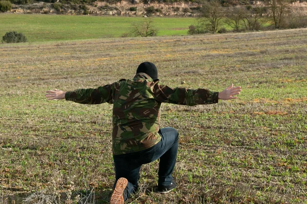 Military man in jeans and balaclava detained with his arms raised in the mountains