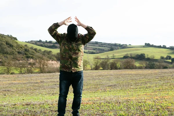 Military man in jeans and balaclava detained with his arms raised in the mountains