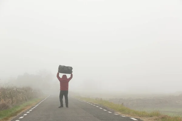 Llevando una maleta en sus brazos un hombre irreconocible caminando por una carretera en un día de niebla Fotos De Stock Sin Royalties Gratis