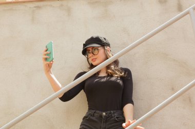 Moda gözlüklü ve şapkalı genç bir kadının güneşli bir günde merdivenlerde durup akıllı telefondan kendi portresini çekmesi.