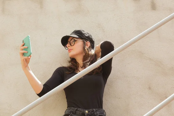 Kendine güveni düşük, şık şapkalı, gözlüklü, saçına dokunan ve yazın gri duvara yaslanırken cep telefonuyla kendi portresini çeken bir kadın..