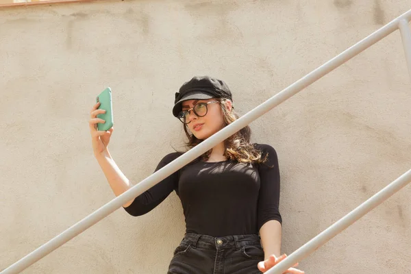 Moda gözlüklü ve şapkalı genç bir kadının güneşli bir günde merdivenlerde durup akıllı telefondan kendi portresini çekmesi.