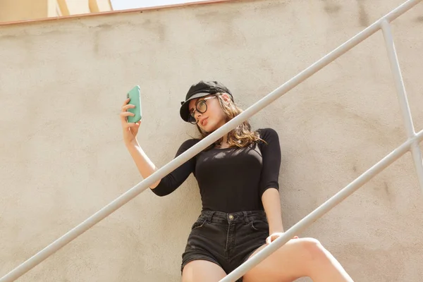 From below of cool young female in black outfit standing on staircase in city and taking selfie on mobile phone in summer sunny day