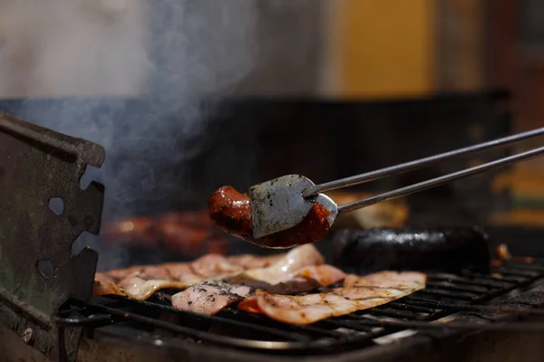 Metal tongs moving smoked pork bacon on outdoor barbecue outside the house. Family Food Day with outdoor barbecue activity.