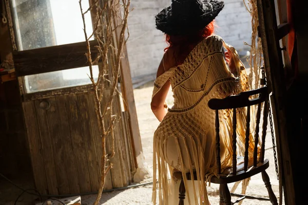 Woman backlit in doorway sitting on chair with hat in old industrial building