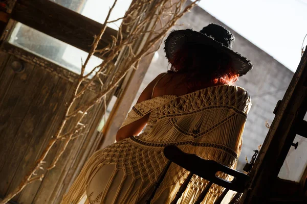 Woman backlit in doorway sitting on chair with hat in old industrial building