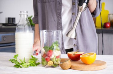 Meyve suyu yapmak için el blenderi kullanan bir kadın. Vejetaryen, temiz yeme tarzı konsepti. Gri mutfak iç tasarımı.