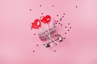 İki kırmızı lolipop şeklinde kalp pembe arka planda küçük alışveriş arabasında Confetti Sevgililer Günü arkaplan alışverişi ya da indirimli tatil konsepti