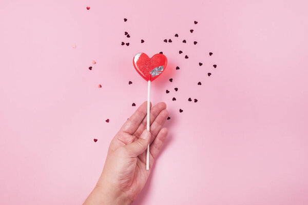 Hand Holding lollipop in Heart Shape Valentines Day Candy Lollypop Confetti Pink Background