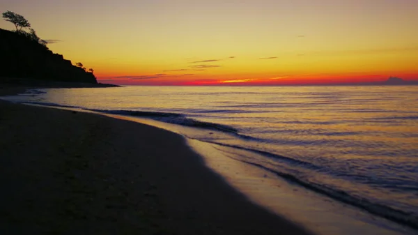 La costa del lago all'alba arancione del tramonto. Spiaggia mare scuro con silhouette di montagna — Foto Stock