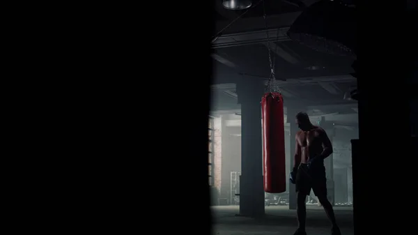 Человек заканчивает тренировку по боксу. Боксер ходит по чердачному зданию с проколотой сумкой — стоковое фото