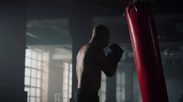 Boxeador golpeando saco de boxeo durante el entrenamiento intenso. Hombre practicando kickboxing en gimnasio —  Fotos de Stock