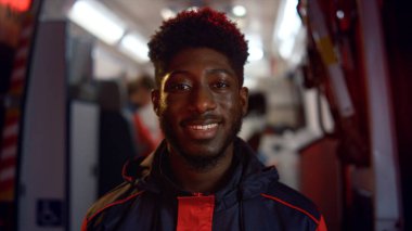 Sağlık görevlisi kameraya gülümsüyor. Erkek doktor, acil durum arabasına karşı duruyor.