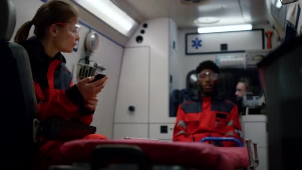 Paramédicos sentados en el coche de emergencia. Médico femenino enfocado usando smartphone — Vídeo de stock