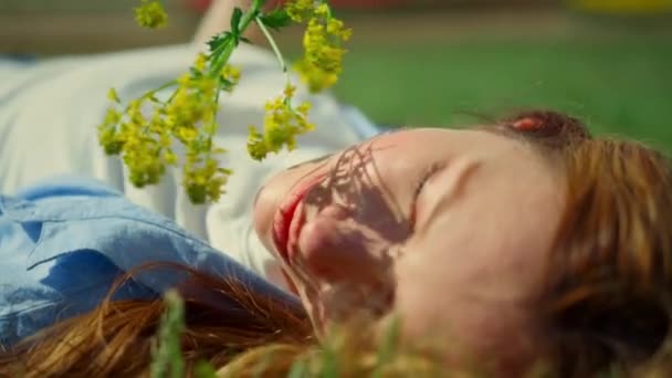 Zrelaksowana dziewczyna pozująca z dzikim kwiatem w zielonym parku. Cień gałęzi na ładnej twarzy — Wideo stockowe
