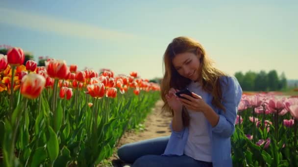 Szczęśliwa kobieta dotykając ekranu telefonu komórkowego podczas siedzenia na drodze na polu tulipan — Wideo stockowe