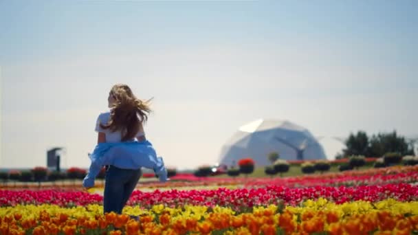 Задний вид смеющейся девушки, бегущей через тюльпановое поле с футуристическим зданием. — стоковое видео