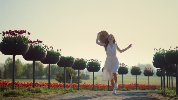 Jonge vrouw rent in het park met bloemen. gelukkig meisje het nemen van uit hoed. — Stockvideo