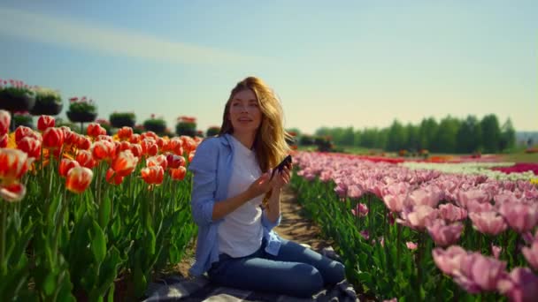 Mladá žena sedí v kvetoucí květinové zahradě s mobilním telefonem ve slunečný den. — Stock video