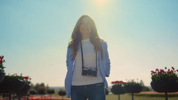 Uśmiechnięta dziewczyna w dżinsach stojąca z kamerą w kwitnącym wiosennym parku w słońcu. — Zdjęcie stockowe