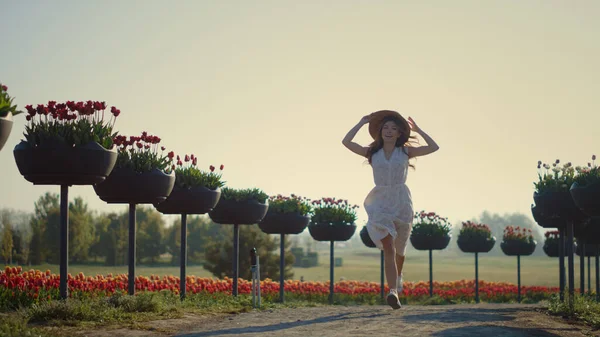 Young woman running in park with flowers. Happy girl taking off hat. — Stock Photo, Image