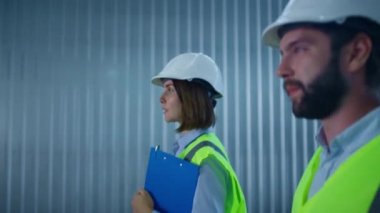 Üniformalı imalat uzmanları depoda dolaşıp ürünü tartışıyor.