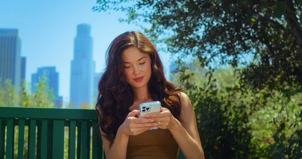Mulher asiática olhando para trás na paisagem urbana borrada segurando smartphone no parque close-up — Fotografia de Stock